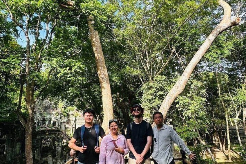 A group of people standing in a forested area with large trees and a stone structure in the background. 2