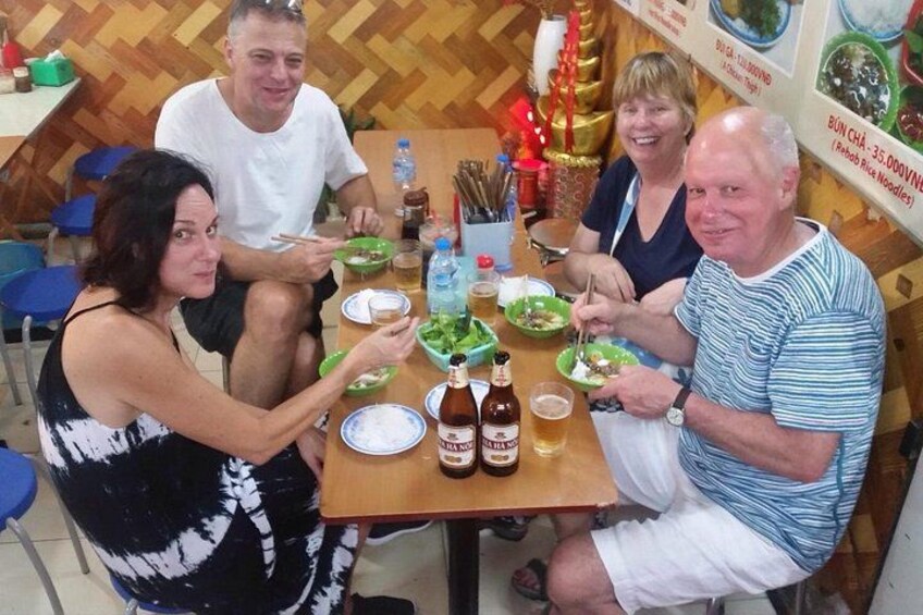 Un grupo de personas sentadas alrededor de una mesa con platos de comida y botellas de cerveza, con una pared decorada al fondo. 1