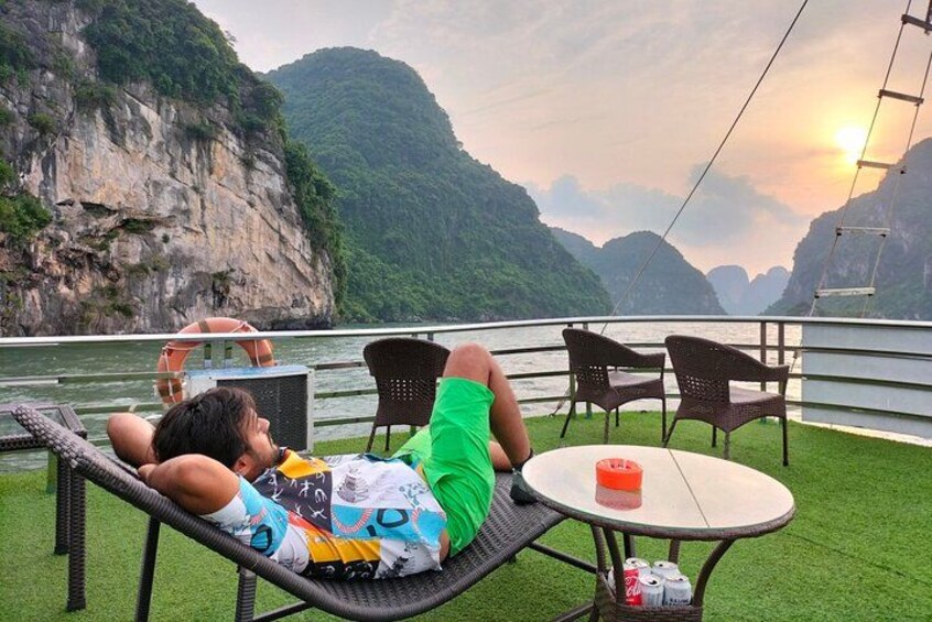 A person relaxing on a lounge chair with a table and chairs, overlooking a body of water and a mountainous landscape during sunset. 4