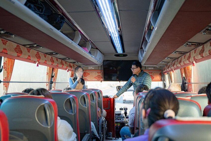 A bus interior with passengers seated, a driver at the front, and a television screen. 2