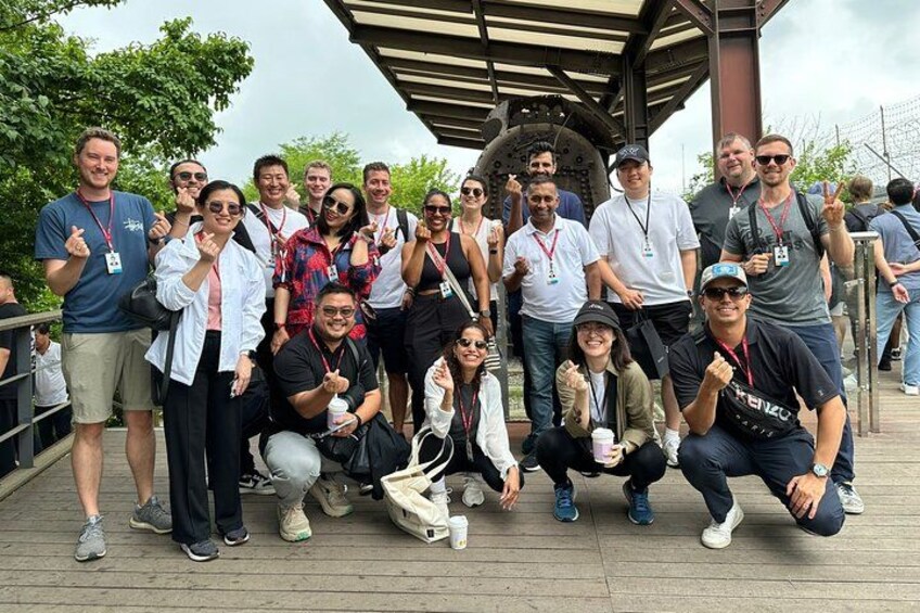 Un grupo de personas posando para una foto en una terraza de madera, con una gran estructura mecánica al fondo. 2