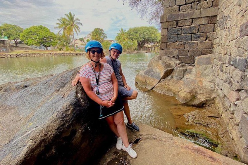 Two people sitting on a rock by a water body, with a stone wall and greenery in the background. 5