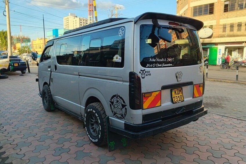A silver van parked on a paved area with a license plate, surrounded by buildings and other vehicles. 2