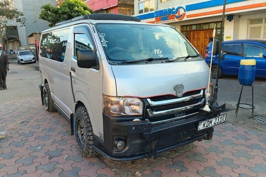 A silver van parked on a paved area with a blue car in the background. 3