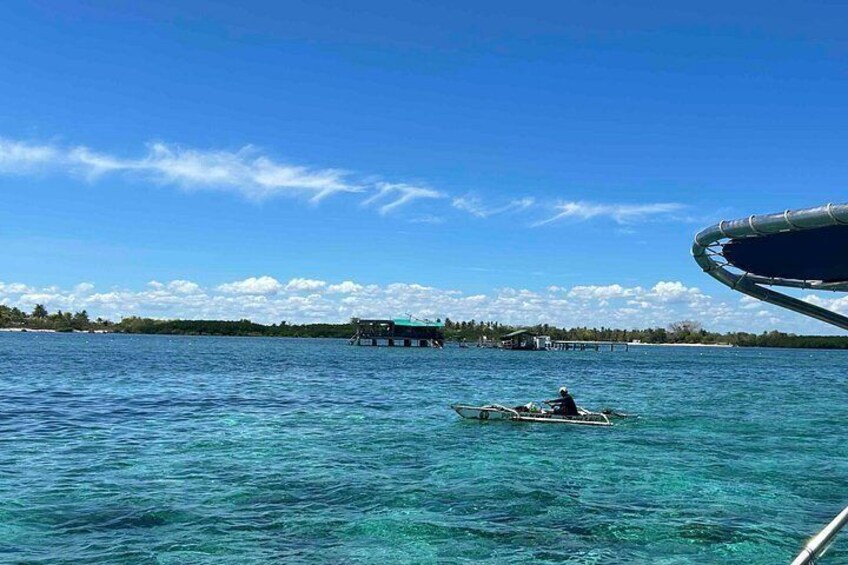 Mar azul y transparente con una persona remando en un bote pequeño, un muelle con construcciones y una orilla distante con árboles. 2