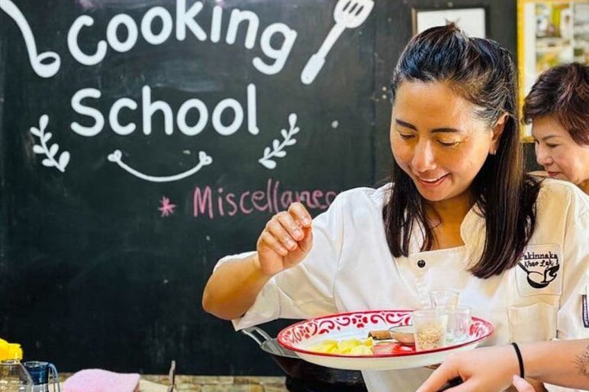 Una persona con uniforme blanco de chef sostiene un plato con comida y una bebida frente a una pizarra con la frase 'Cooking School' escrita. 5