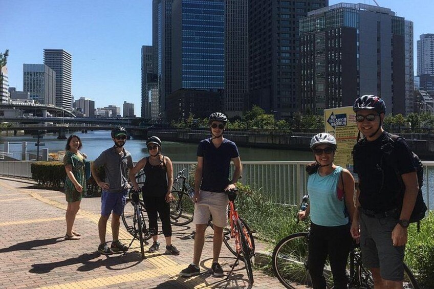 Un groupe de personnes se tient debout avec des vélos dans un environnement urbain, avec une rivière et des bâtiments en arrière-plan. 2