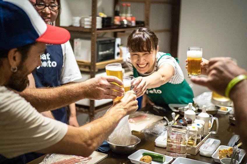 Eine Gruppe von Personen stöstet mit Biergläsern an einem Tisch an, auf dem verschiedene Speisen und Küchenutensilien stehen. 1