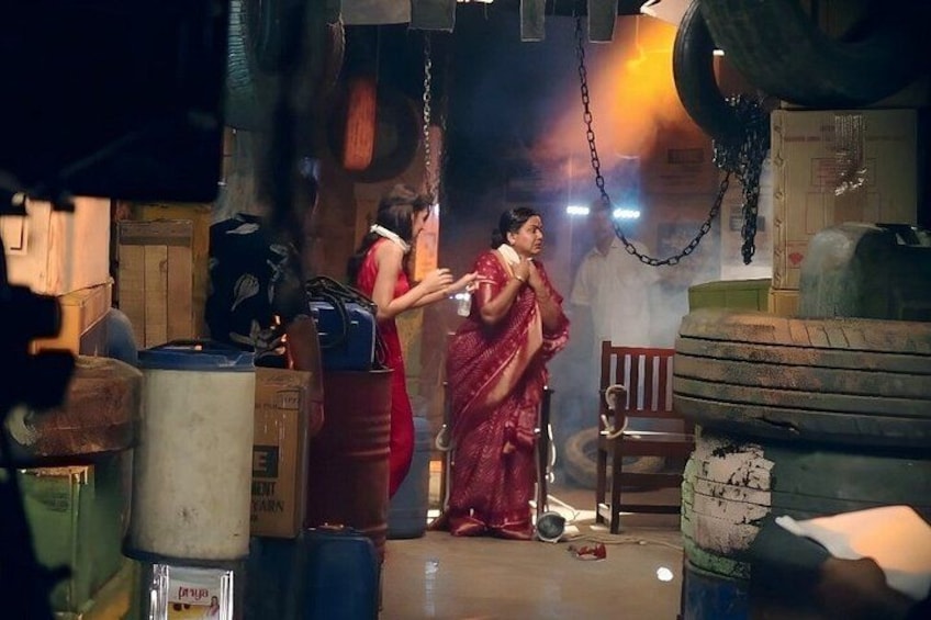 A woman in a red saree standing in a cluttered room with various objects and a large container. 1