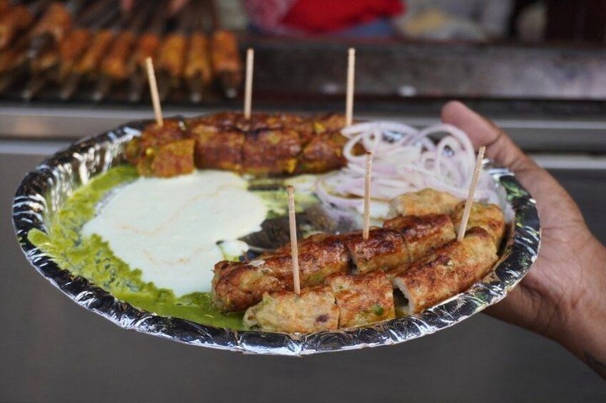 A hand holding a foil plate with grilled kebabs, a dollop of white sauce, and sliced onions. 4
