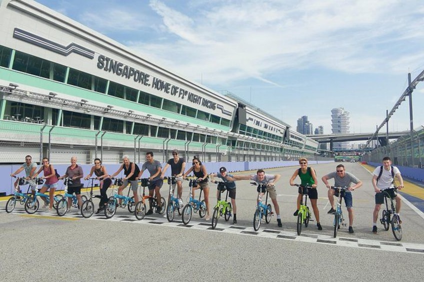 En gruppe cyklister foran en moderne bygning med teksten 'SINGAPORE, HOME OF F1 NIGHT RACING'. 5