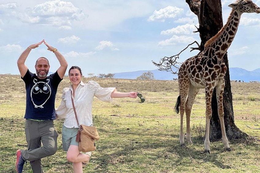 Un hombre y una mujer posando con una jirafa en un campo cubierto de hierba, con montañas al fondo. 3