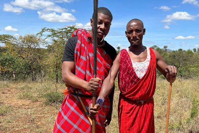 Two individuals in traditional attire standing in a natural landscape with trees and a clear sky. 3