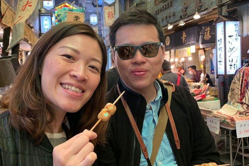 A person holding a skewer with food, another person in the background, and various signs and stalls. 5