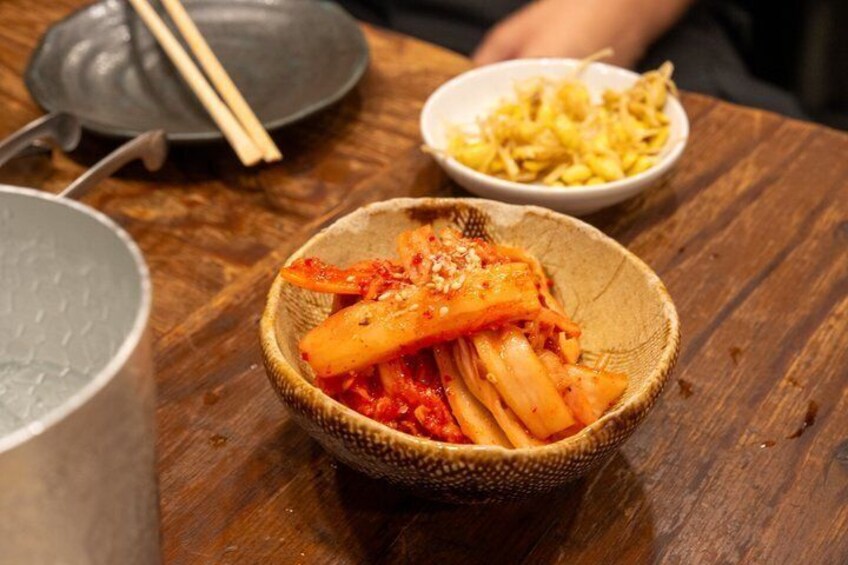 Et træbord med kimchi, syltede grøntsager, spisepinde og en gryde. 2