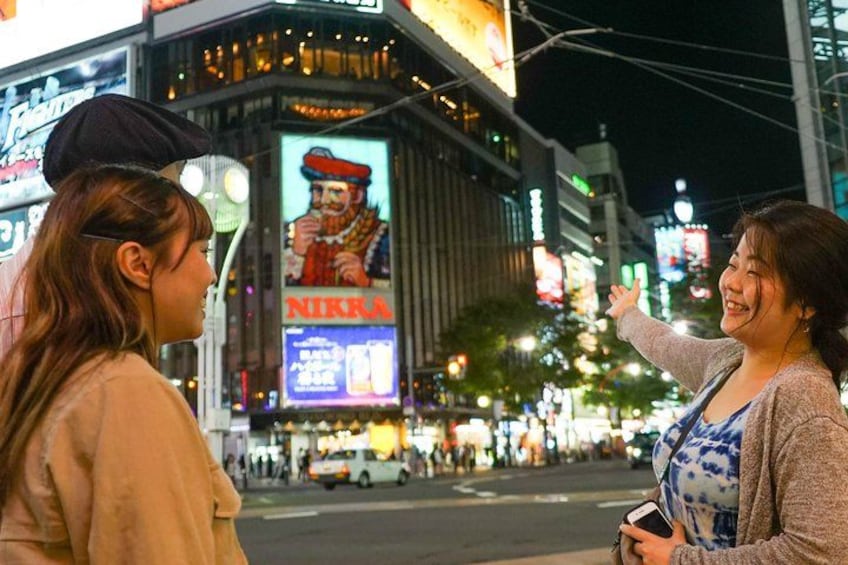 Dos personas conversando en una calle urbana de noche, con vallas publicitarias iluminadas al fondo. 2