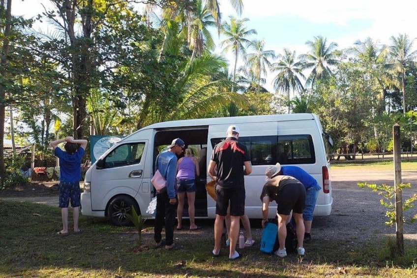 A group of people around a white van in a grassy area with palm trees in the background. 3