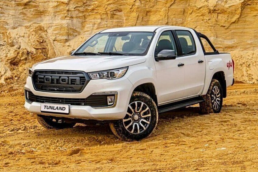 Een witte FOTON pick-up truck met 'TUNLAND' op het kenteken, geparkeerd op een zandgrond oppervlak met een rotsachtige achtergrond. 3