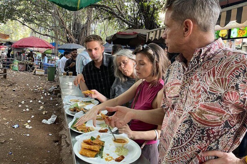 People eating at an outdoor food stall with tables and food remnants on the ground. 5