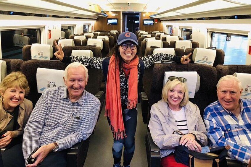 A group of people seated in a train carriage with overhead luggage racks and personal space. 2