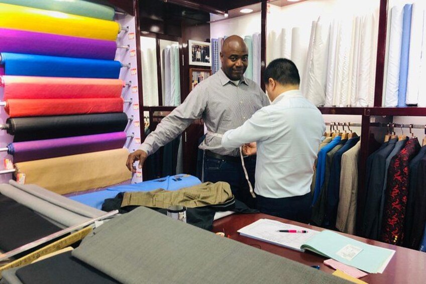 A tailoring shop with colorful fabric rolls, a mannequin, and a man measuring a suit. 2