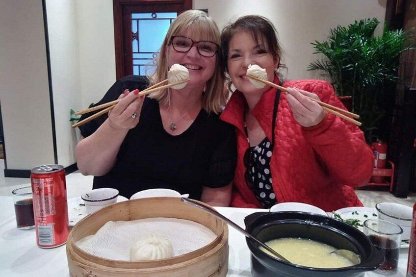 Two people eating dumplings with chopsticks, a bamboo steamer, a pot of soup, and a can of soda. 4