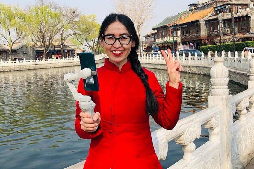 A person in a red dress holding a smartphone with a selfie stick, standing by a body of water with traditional architecture in the background. 2