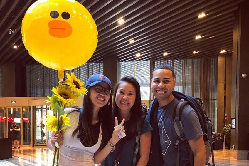 Three people in an indoor setting with a large yellow balloon, a sunflower bouquet, and a modern interior with a sloped ceiling and large windows. 4