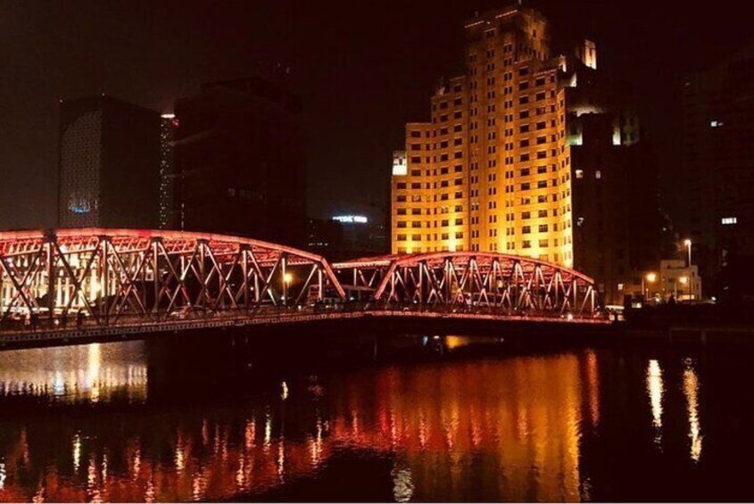 A lit bridge at night with reflections on the water, a tall building in the background, and city lights. 3