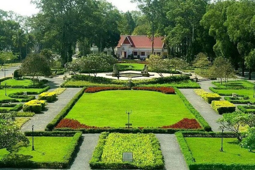 En välskött trädgård med geometriskt utformade blomsterrabatter, en central gräsmatta och en byggnad med ett rött tak i bakgrunden. 4
