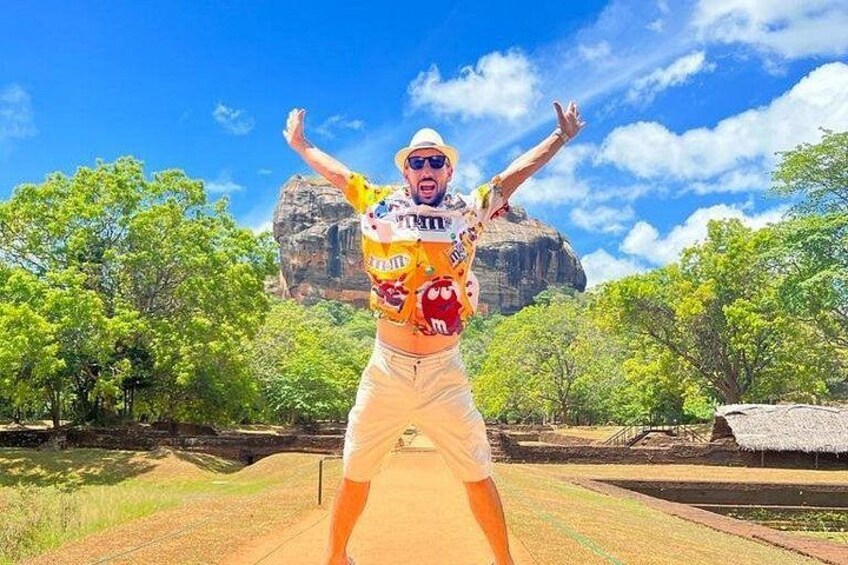 Une personne portant une chemise et un short colorés, debout dans un environnement naturel, avec des arbres et une grande formation rocheuse en arrière-plan. 1