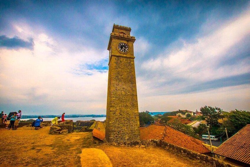 Een stenen klokkentoren met wijzerplaat, omringd door een historisch fort en een kustlandschap. 2
