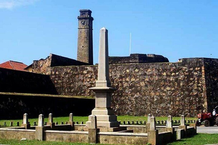 En stenvæg med et tårn med et ur, et monument og et fortorv med et hegn. 2
