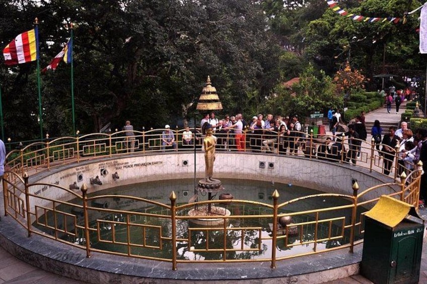 Una fontana circolare con una statua al centro, circondata da un parapetto. Si vedono delle bandiere sullo sfondo. 5