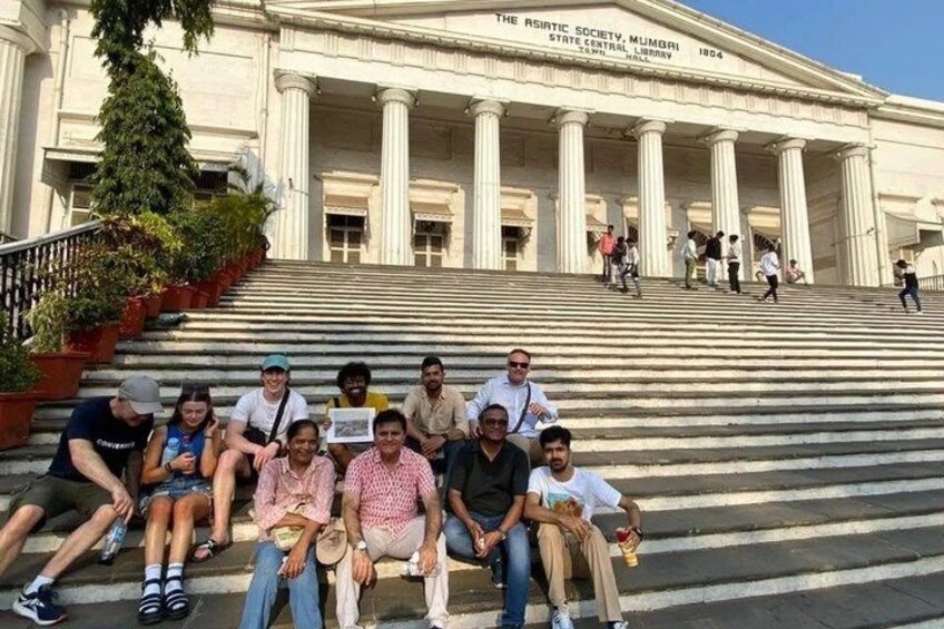Un grupo de personas sentadas en escalones frente a un edificio con columnas y un letrero que dice 'The Asiatic Society, Mumbai'. 4