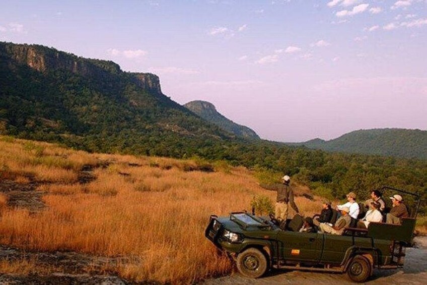 A group of people in a green vehicle on a grassy field with mountains in the background. 1