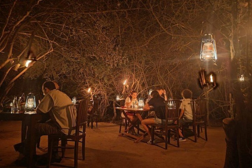 An outdoor dining area at night with tables and chairs, surrounded by trees and illuminated by lanterns. 2