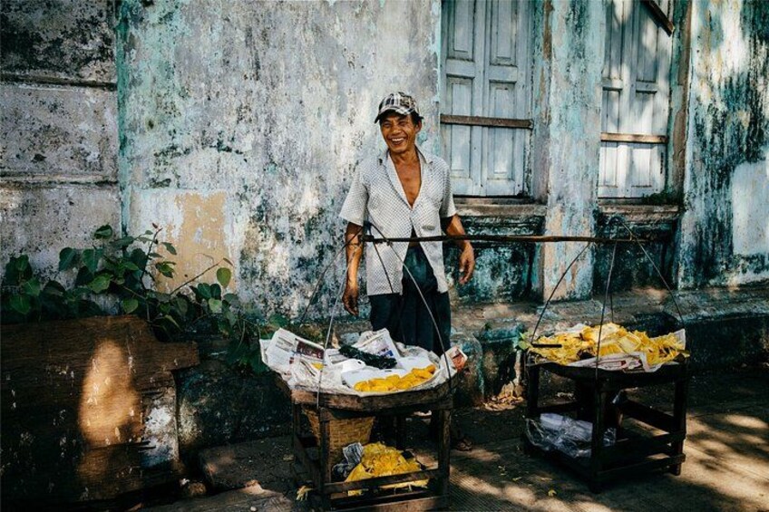 A person standing next to a cart with newspapers and yellow items, in front of a weathered building with a window. 4