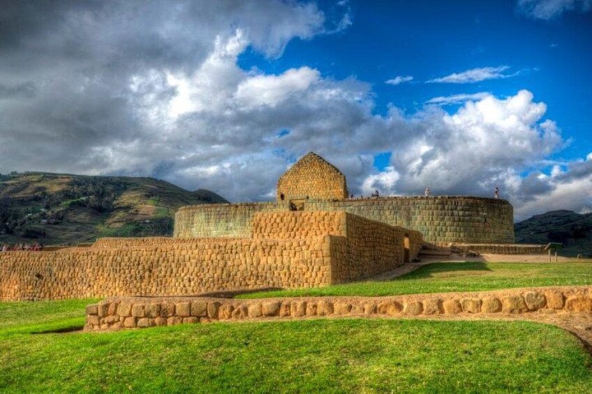Una fortaleza de piedra histórica con una torre central destacada, rodeada de un área verde bien cuidada y un telón de fondo montañoso. 2