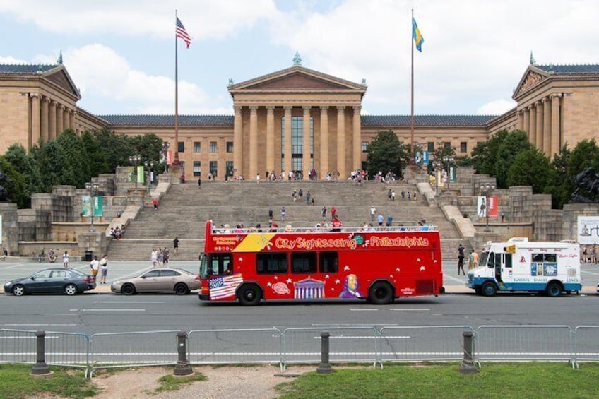 A red bus with 'Cuyahoga Valley' and 'Philadelphia' written on it, parked in front of a large building with steps leading up to it. 2