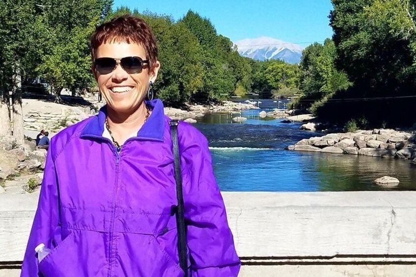 A person in a purple jacket standing by a river with rocks and trees in the background. 3