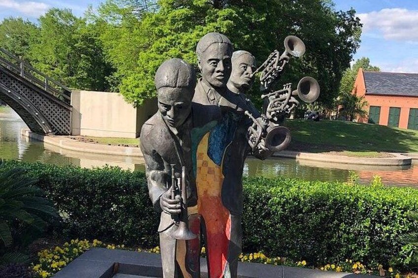 A statue of three figures, one holding a trumpet, set against a backdrop of a bridge, water, and greenery. 3