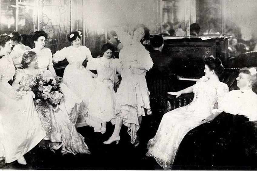 Een groep mensen in retro kleding, sommigen zitten en sommigen staan, in een kamer met een piano. 5