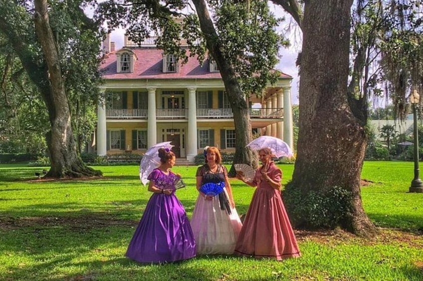 Three women in vintage dresses standing in a grassy area with a large house in the background. 3