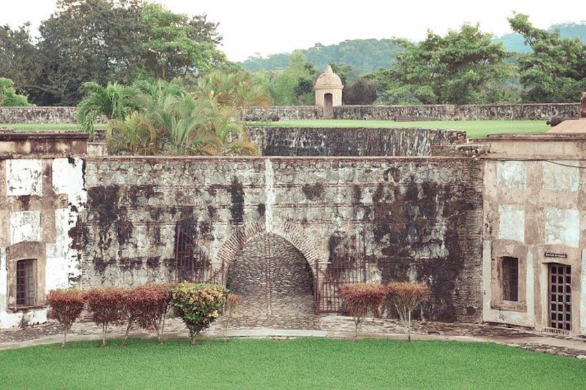 A historic stone wall with an arched gateway, surrounded by well-maintained shrubbery and a small building in the background. 1
