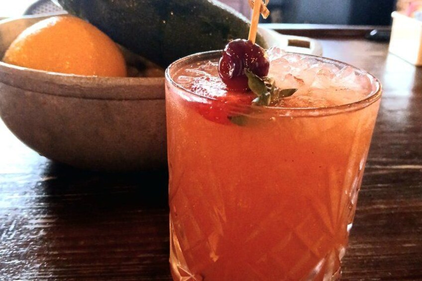 Un vaso de bebida roja con cereza y una hoja de adorno, un cuenco de fruta y una mesa de madera. 3