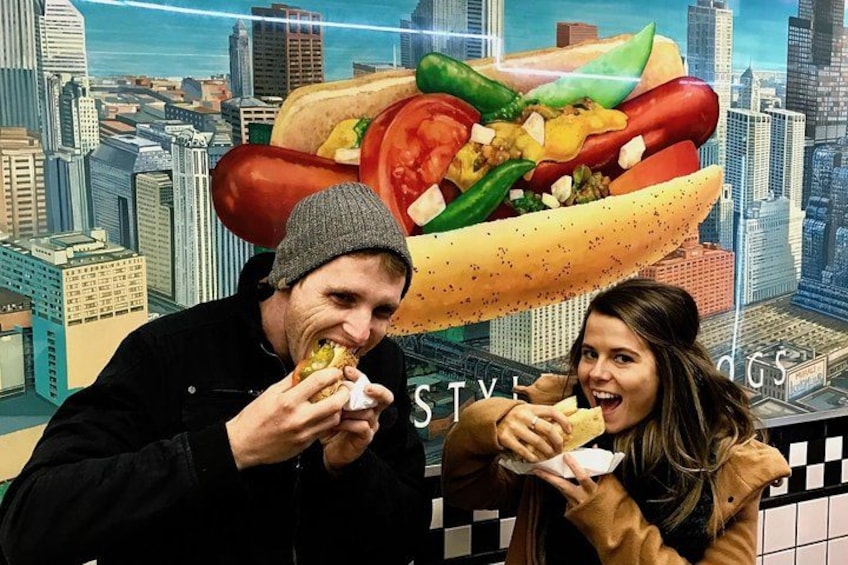 Two people eating hot dogs in front of a cityscape backdrop. 1