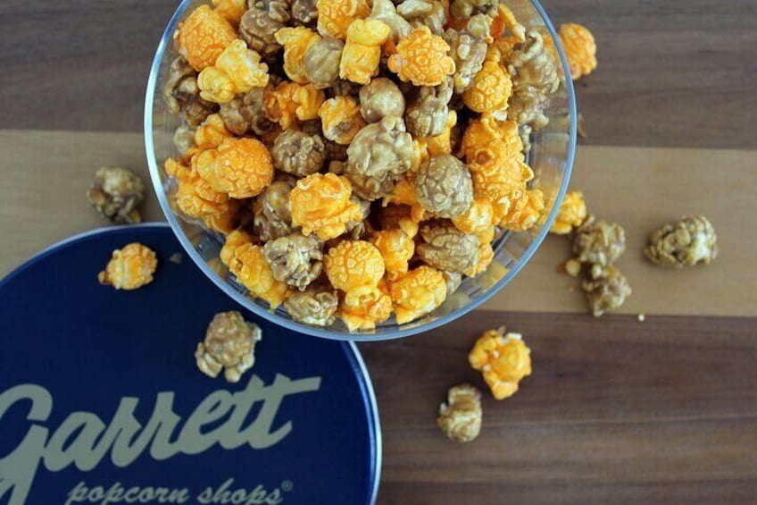 Un tazón de palomitas de maíz con tonos anaranjados y marrones, un envase azul con el texto 'Garrett popcorn shops' y palomitas esparcidas sobre una superficie de madera. 5