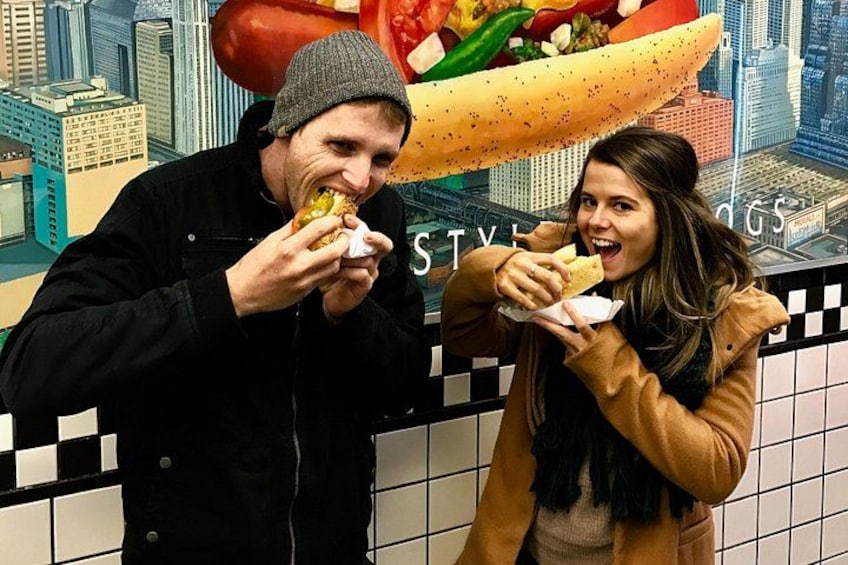 Two people eating hot dogs in front of a cityscape backdrop. 2