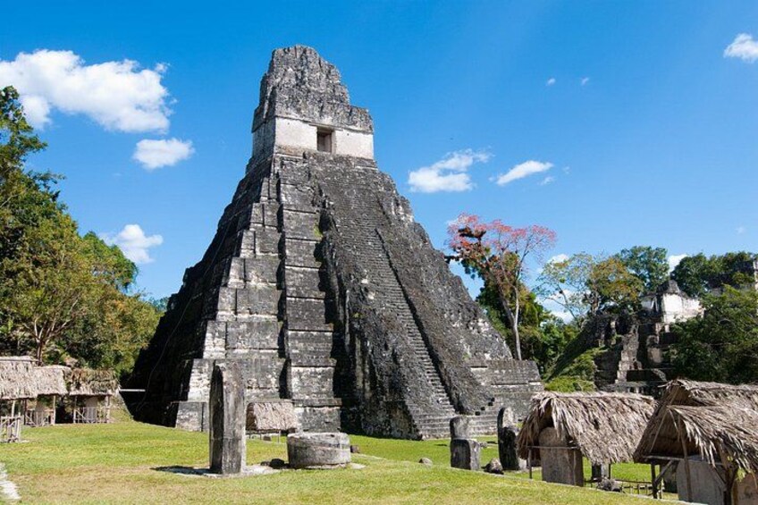 Una gran estructura en forma de pirámide con un edificio más pequeño en la parte superior, rodeada de árboles y chozas con techo de paja. 3